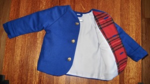 Blue raglan sleeve coat with gold buttons with right side flipped open to reveal white satin lining, red plaid facing, and blue hanging loop
